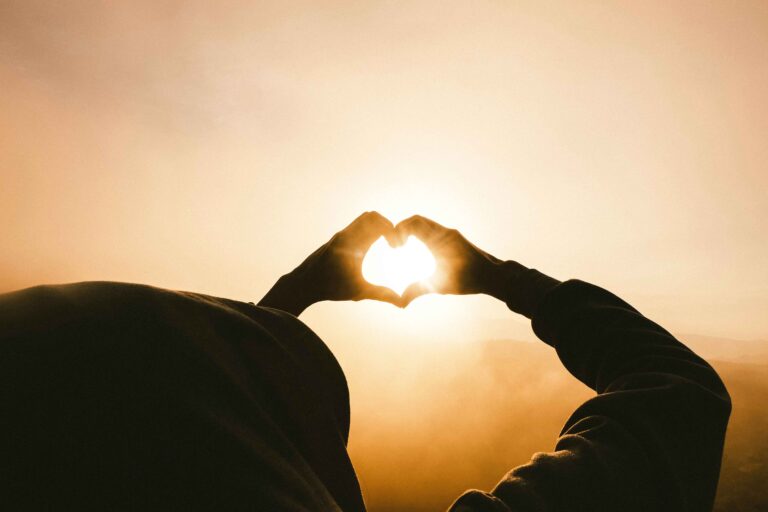 A person holding their hands up in a heart shape with the sun in the background