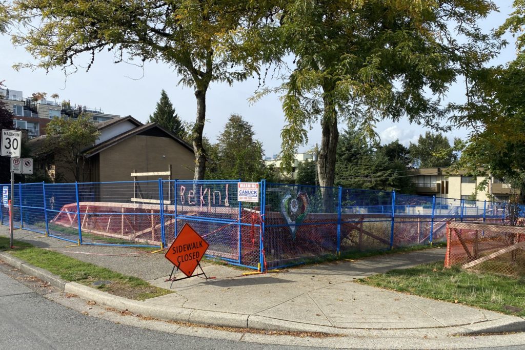 Corner street view showing a demolished lot surrounded by blue metal fencing with "Be kind" message taped on one side
