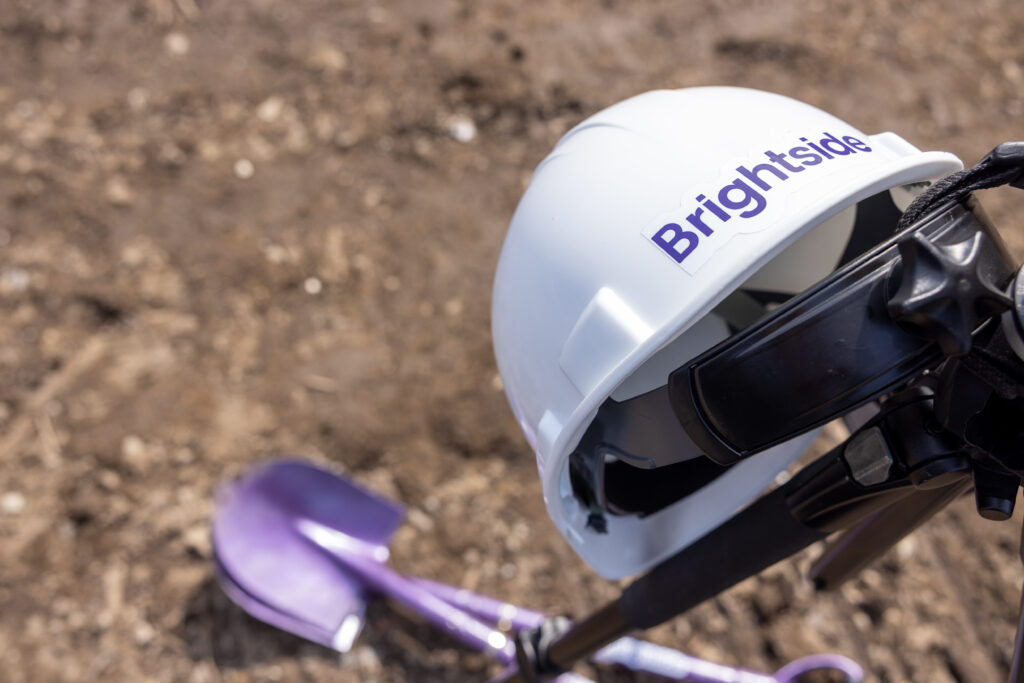 Photo of a Brightside-branded hard hat resting on a tripod on the dirt ground, with two out-of-focus purple shovels lying at the base of the tripod.